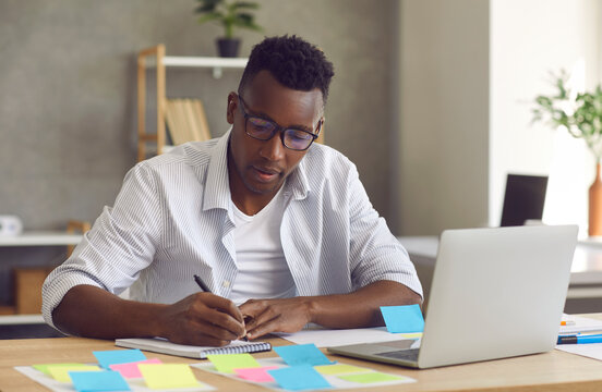 Startup project development, research work, brainstorm. Young handsome minded black african american businessman sitting at home table front of laptop writing down ideas in notebook