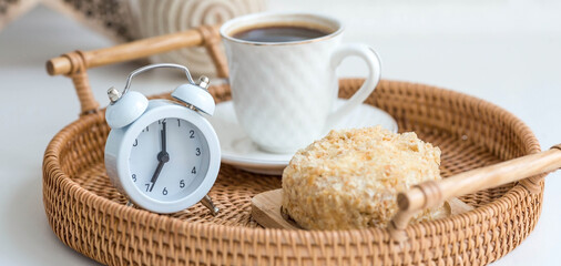 A white retro alarm clock, a cake and a cup of coffee against a white brick wall. The concept of...