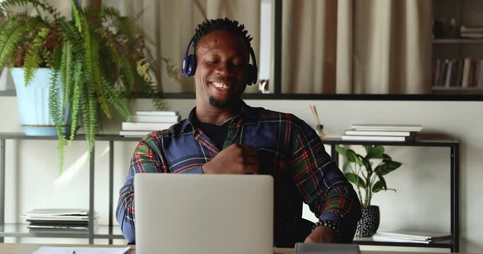 Relaxed young happy african ethnicity biracial male employee in headphones enjoying pleasant video conference zoom conversation with colleagues, discussing project or working issues in office.