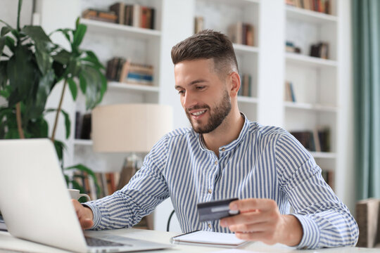 Smiling man sitting in office and pays by credit card with his laptop.