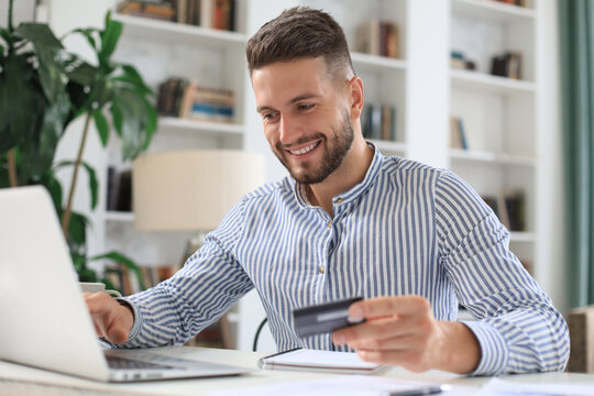 Smiling Man Sitting In Office And Pays By Credit Card With His Laptop.