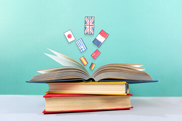 On the table is a stack of books, with an open book on top, from which flags fly out. Turquoise background. The concept of learning foreign languages