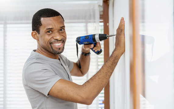 Portrait Of Happy African American Black Man Using Electric Drill On Wall. DIY, Do It Yourself Hobby, Handyman Worker Using Drill While Working On Construction Site. Home Interior Builder Moving 