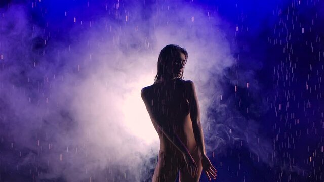 Silhouette of young beautiful ballerina woman emotionally dancing modern contemporary ballet dance under rain and splashing water. Shot in dark studio with smoke and blue lights. Slow motion. Close up