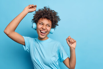 Positive Afro American girl enjoys good day keeps arms raised has fun poses carefree wears...