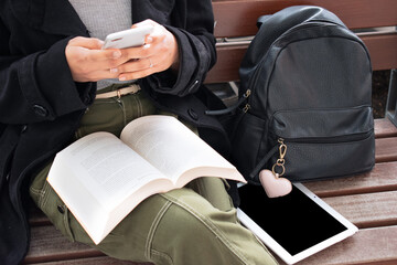 close up of a black girl with no identity sitting on a wooden bench using a cell phone with an open book resting on her legs, a backpack and a tablet on one side.