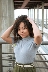 young black woman in gray t-shirt and green pants looking at the camera while touching her hair.