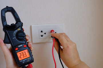 An electrician is a hand holding on the digital multimeter into a hole of an electric socket to ...