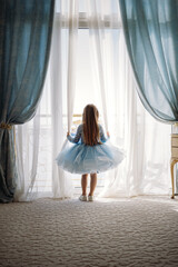 Cute little Caucasian girl in babydoll dress standing by big french window in bedroom looking outside at the sea. Image with selective focus