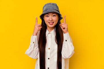 Young chinese woman isolated on yellow background indicates with both fore fingers up showing a...