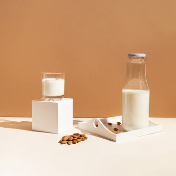 Glass with vegan milk from almond nuts with a bottle on white wooden tray with sunlit background. Dairy and lactose free substitute drink. Healthy food ingredient concept. Trendy colors.
