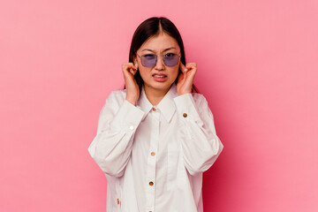 Young chinese woman isolated on pink background covering ears with fingers, stressed and desperate by a loudly ambient.