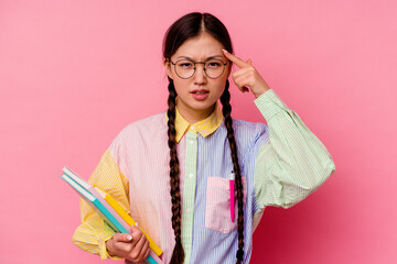 Young chinese student woman holding books wearing a fashion multicolour shirt and braid, isolated on pink background showing a disappointment gesture with forefinger.