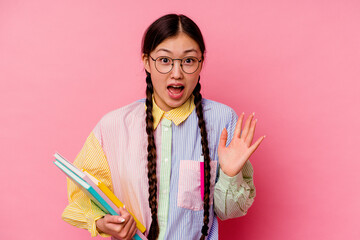 Young chinese student woman holding books wearing a fashion multicolour shirt and braid, isolated on pink background receiving a pleasant surprise, excited and raising hands.