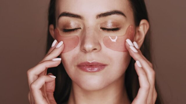 Skin care. Facial treatment. Skin moisturizing rejuvenation. Beauty portrait of peaceful relaxed woman with nude makeup applying pink collagen gel eye patches isolated on brown.