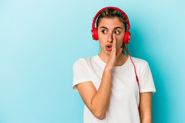 Young blonde woman listening to music on headphones isolated on blue background is saying a secret hot braking news and looking aside