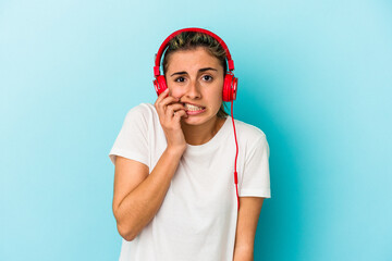 Young blonde woman listening to music on headphones isolated on blue background biting fingernails, nervous and very anxious.