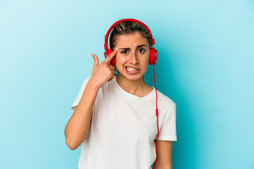 Young blonde woman listening to music on headphones isolated on blue background showing a disappointment gesture with forefinger.