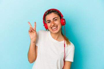 Fototapeta premium Young blonde woman listening to music on headphones isolated on blue background joyful and carefree showing a peace symbol with fingers.