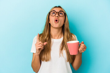 Young caucasian blonde woman holding a mug isolated on blue background pointing upside with opened mouth.