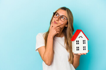 Young caucasian woman holding a house model isolated on blue background looking sideways with...