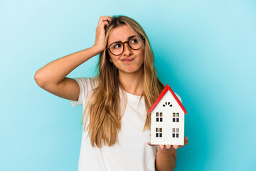 Young caucasian woman holding a house model isolated on blue background being shocked, she has...