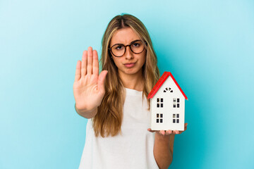 Young caucasian woman holding a house model isolated on blue background standing with outstretched...