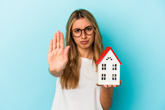 Young caucasian woman holding a house model isolated on blue background standing with outstretched hand showing stop sign, preventing you.