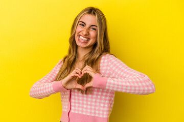 Young blonde caucasian woman isolated on yellow background smiling and showing a heart shape with hands.