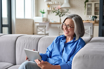 Happy middle aged senior woman sitting on couch holding using digital tablet device during video call with family friends. Older female reading ebook or watching zoom video meeting online.