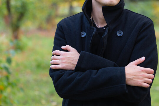 a man stands in a black coat with his arms folded on his chest expressing feelings of concern and anxiety against the background of a green park copy space close-up