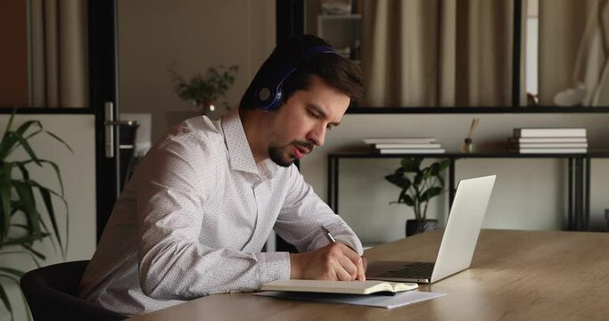 Focused young happy hispanic businessman manager in headphones watching educational video on computer, writing notes in copybook, improving professional knowledge distantly or attending virtual event.