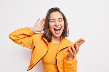 Overjoyed pretty Asian teenage girl got carried away with music holds smartphone enjoys favorite...