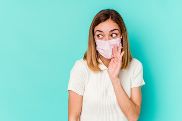 Young woman wearing a mask for virus isolated on blue background is saying a secret hot braking...