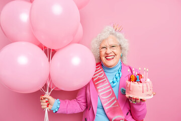 Fototapeta na wymiar Life only start when you get older. Glad wrinkled old woman celebrates birthday holds festive cake with burning candled bunch of inflated balloons isolated over pink background. Party for old people