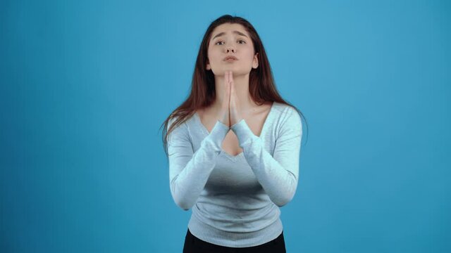 The desperate young woman manifests her prayer by displaying her hands in the form of a prayer. Asian with dark hair, dressed in a blue blouse, isolated on a dark blue background in the studio. The