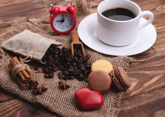 white cup of black coffee, heart, beans, macaroons, cinnamon and clock on the wooden background