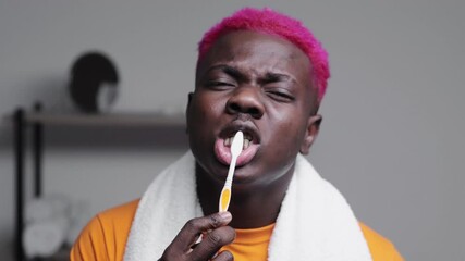 Morning hygiene. Stylish black man. Bathroom routine. Healthy treatment. Smiling hipster african guy bright pink hair towel on shoulder brushing teeth gray interior background.