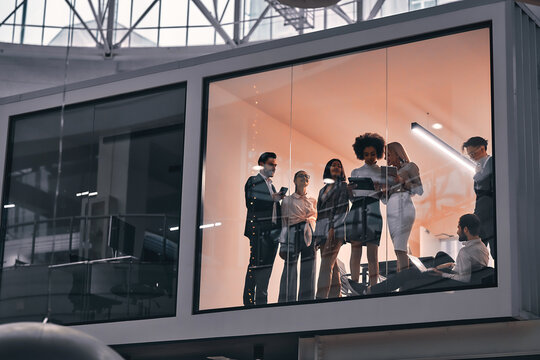 Behind The Glass In The Meeting Room In The Spacious Large Business Center, A Team Of Young People Stand And Discuss The Work.