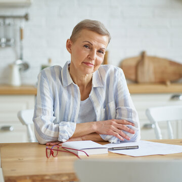 Home Office. Portrait Of Beautiful Middle Aged Woman Smiling At Camera While Sitting At Wooden Table In Kitchen And Working With Documents. Senior People And Distance Work Concept