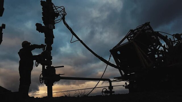 An employee in overalls and a helmet is carrying out repairs and maintenance of an oil well. Silhouette on the background of the evening sky