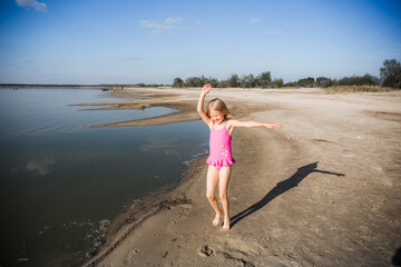 A little seven-year-old girl in a pink swimsuit runs along the lake in a good cheerful mood with a smile on her face. Summer lifestyle. Health tempering with water procedures and outdoor walks.