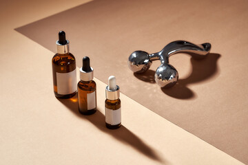 Three glass eye dropper serum bottles and modern metal 3D face lift roller isolated on beige surface