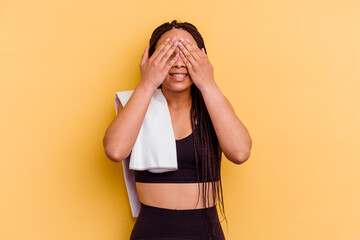 Young sport african american woman holding a towel isolated on yellow background covers eyes with hands, smiles broadly waiting for a surprise.