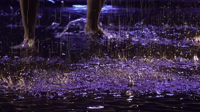 Contemporary dance ballerina dances sensually in rain on water surface, creating splashes. Silhouette of wet woman in white shirt move plastically in dark studio with blue light. Slow motion. Close up