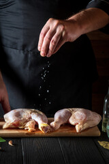 The cook adds salt to the chicken legs. Preparation for grilling with chicken leg in the kitchen of a restaurant or cafe on table with vegetables and spices