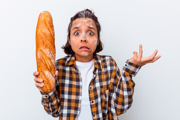 Young mixed race woman making bread isolated on white background shrugs shoulders and open eyes...