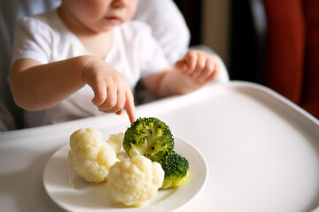 a toddler sits behind a high chair and points at the broccoli. Concept: proper nutrition for...