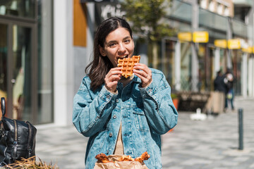 cheerful young woman enjoying tasty waffles in front of a cafe in the street. take away food in the...