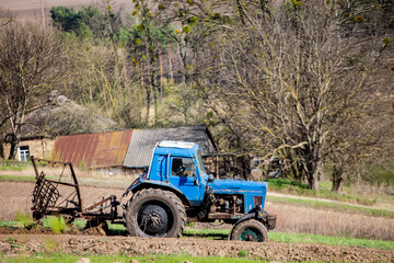 Naklejka premium old blue tractor with plow on field and cultivates soil. Preparing the soil for planting vegetables in spring. Agricultural machinery, field work.